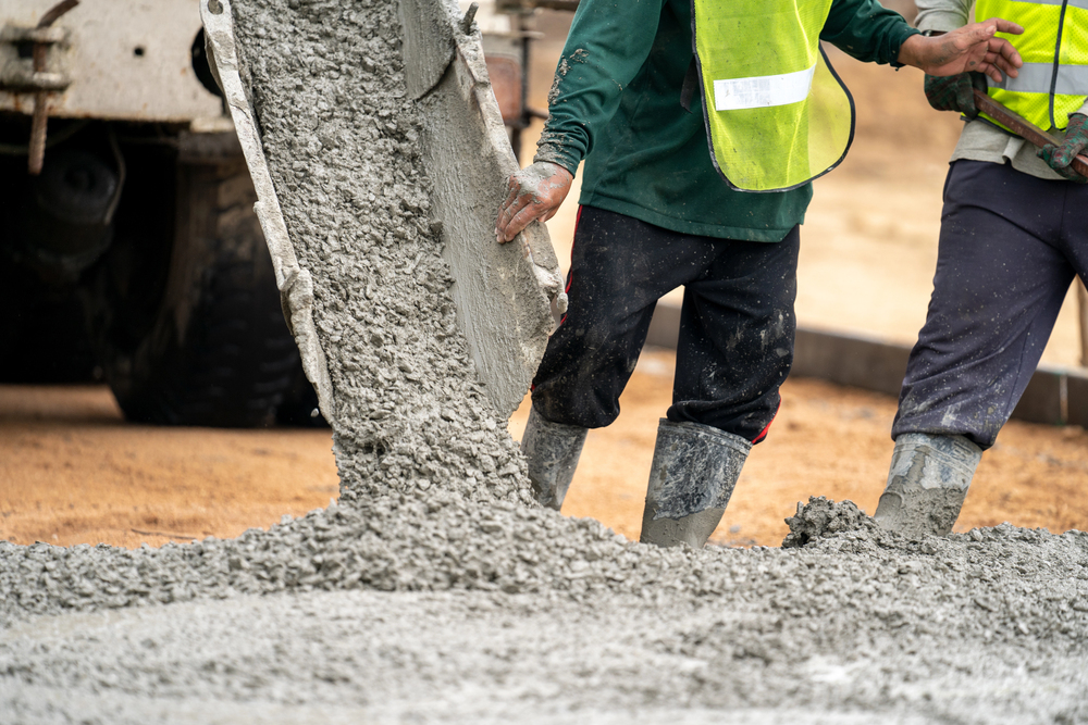 Construction workers in safety vests pouring and directing fresh concrete from a mixer truck chute at a job site.
