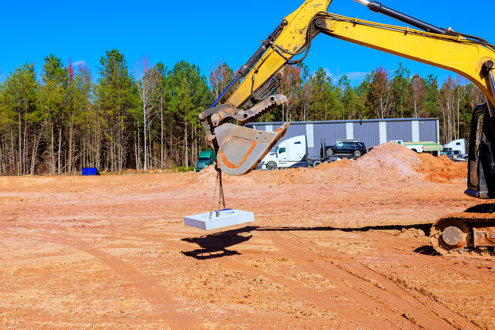 Yellow excavator lifting a concrete slab with chains at a construction site with red clay soil and trees in background.